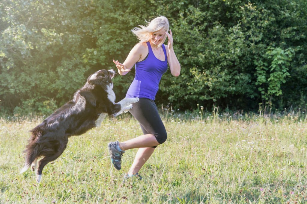 Young blond woman is running with her dog outdoors in the sunny day